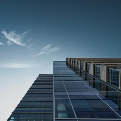 View of the side of a tall glass building looking up to the blue sky with the sun shining in the background